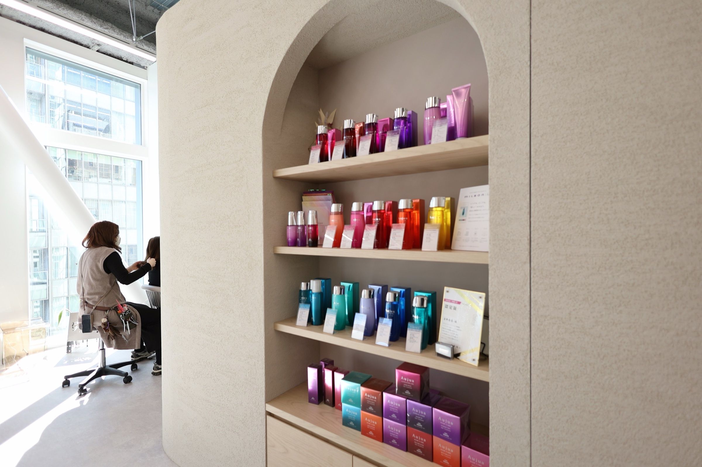 アーチ型の棚に並ぶAujua（オージュア）のヘアケア商品。奥の窓際でスタイリストが施術している